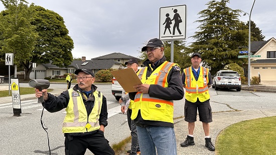 Photo de quatre bénévoles vêtus de gilets de haute visibilité et d'un policier en uniforme effectuant une surveillance de la vitesse dans une zone scolaire.