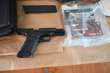 Une arme de poing noire, un chargeur de cartouches et deux boîtes de munitions dans un sac transparent sont étalés sur une table.
