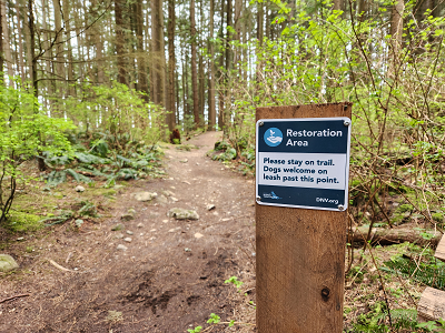 Picture of a wooden post in a wooded area with a sign that states "Restoration area, Please stay on trail. Dogs welcome on leah past this point." 