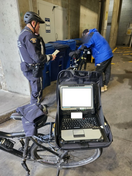 Policier debout à côté de sa bicyclette avec un ordinateur portable professionnel ouvert. Il parle dans sa radio et regarde un homme vêtu d’une veste bleue et d’un pantalon noir qui s’appuie contre un vélo de couleur noire