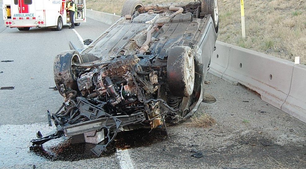 Photo of an upside down crashed car in front of a BC Ambulance on the road.