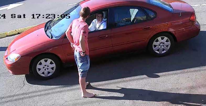 Le suspect se tient à côté d’un petit véhicule orange ou rouge à quatre portes et semble parler au conducteur. Le conducteur est un homme ayant de courts cheveux foncés et portant un tee-shirt blanc ainsi que des lunettes à monture foncée.