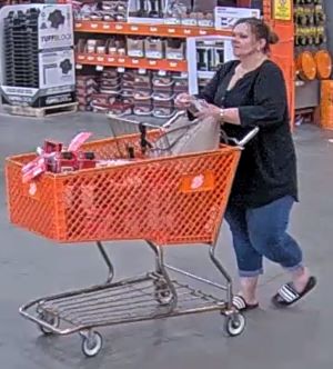 heavy set woman pushing a Home Depot Shopping Cart at the exit of Home Depot. Her cart is now full of tools. She has long brown hair which has been placed into a bun and is wearing blue jeans, a long sleeve black button up shirt, and black sandals with white stripes