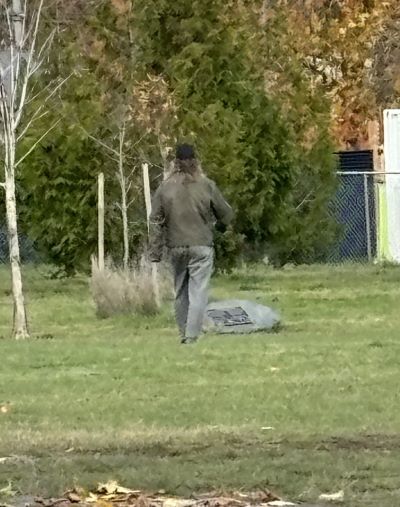 man walking away in a field. He has long grey / brown hair and is wearing a black hat with a light brown/grey leather jacket and grey pants