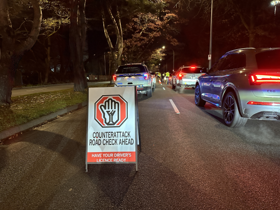 Panneau sur la route qui affiche le message « Contrôles routiers dans le cadre de la campagne Contre-attaque : préparez-vous à présenter votre permis de conduire ». On voit un véhicule de police et des voitures en arrière-plan.