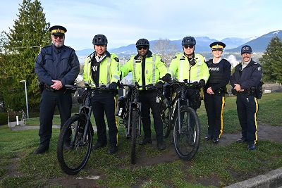 Six police officers wearing various uniforms stand on a grassy area with mountains in the background. Three of the officers have E-bikes in front of them.