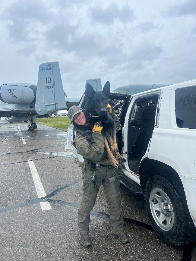 Female police officer, in green uniform carrying her police dog on her shoulder. They are standing in front of a white SUV on a tarmac. 