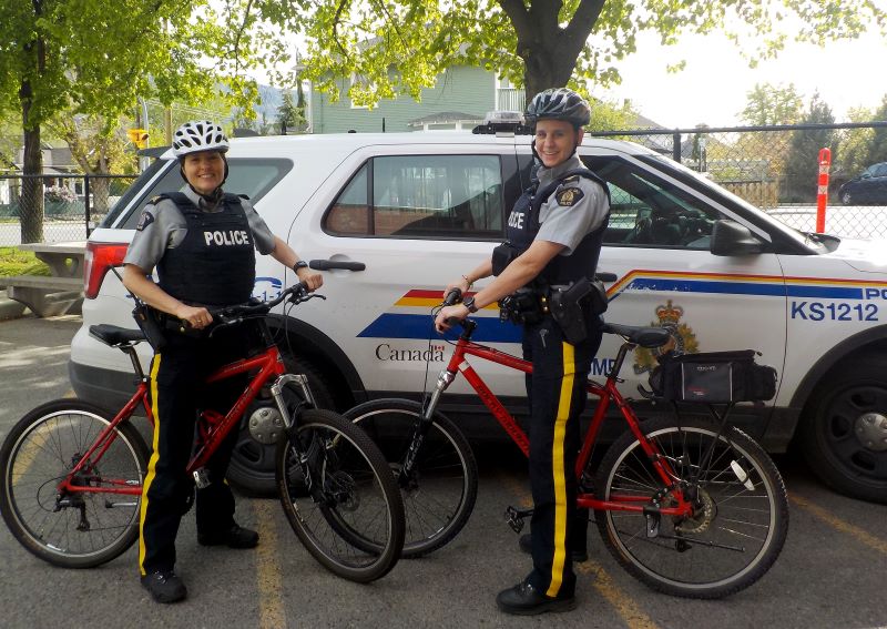 Deux policières portant des casques de vélo sur des vélos de police rouges dans un stationnement. Un VUS de la police est visible derrière elles. 