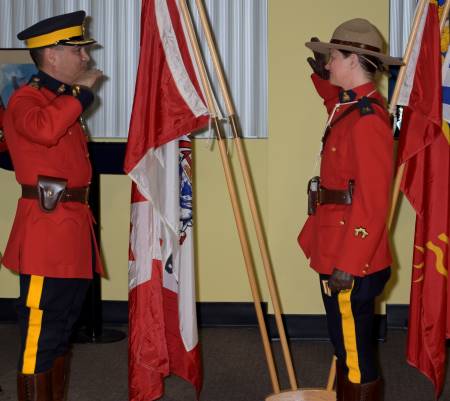 Le surintendant Syd Lecky présente à la gendarme Crystal Evelyn le grade de caporal lors d’une cérémonie plus tôt cet été. À gauche, un homme en tunique rouge salue une femme en tunique rouge. Les drapeaux du Canada et de la Colombie Britannique sont à l’arrière plan. 
