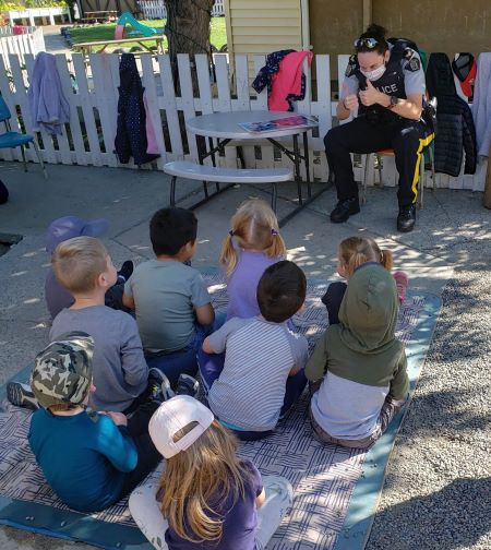 Une photo de la cap. Napier lors d’une discussion sur la sécurité avec des enfants. (Les enfants sont assis en groupe devant une policière en uniforme, qui lève le pouce en l’air. Les manteaux des enfants sont suspendus sur une clôture en bois blanche derrière la policière.) 