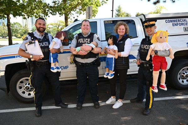 Three smiling RCMP officers pose while holding mannequins beside a BCAA employee in front of an RCMP truck on a sunny day during a child car seat initiative