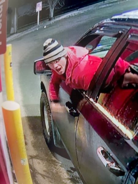 A Caucasian man in a red coat, glasses, and a black and white horizontal striped toque, has his head out the driver’s side window of a vehicle. He appears to be yelling at a vehicle behind him, well stopped at a drive-thru window with a yellow pillar on each side. 