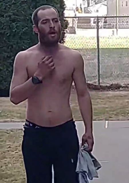 A man with reddish short hair and a beard is standing with his shirt off. His pants are dark. 