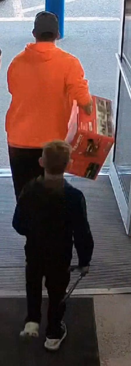 A back image of the orange-sweater suspect carrying a box out of the store. A child in dark clothing is behind him. 