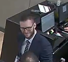 Caucasian man with short light brown hair and short beard, wearing a dark suit jacket with a striped tie, glasses and white dress shirt. The man is facing left in this photo, with a desk behind him.