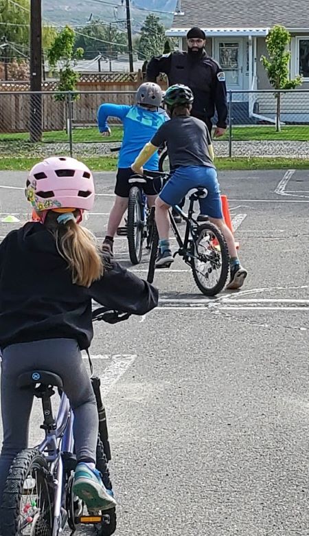 A Kamloops Community Service Officer demonstrates a hand signal to three students. The students are on bikes with helmets. 
