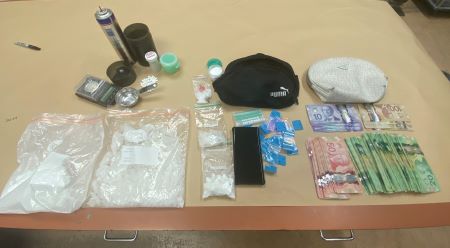 Items seized during a traffic stop are displayed on a table, including bags of white substances, stacks of Canadian currency, pills, a digital scale, two handbags, and other items.  