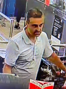 male suspect inside the store. He is wearing a light coloured gold shirt. He has a buzz cut with big dark eyebrows, dark coloured hair and 5 o’clock shadow on his face.