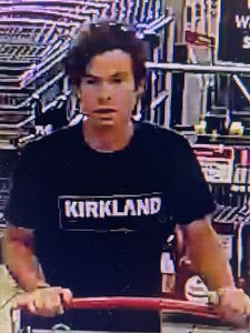 Un suspect pousse un chariot à l’intérieur du magasin. Il porte un t shirt noir orné de l’inscription « Kirkland » en blanc à l’avant. Il a des cheveux bruns courts peignés sur le côté.