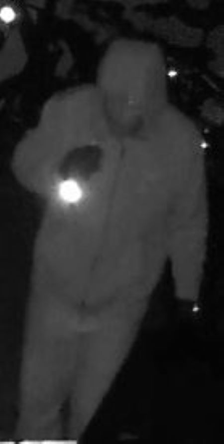 Suspect with flashlight: A suspect in a zipped-up jacket and hoodie, shown in the night vision mode of a security video. 