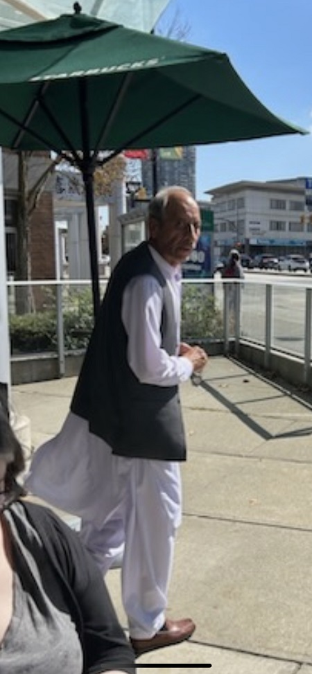South Asian man, balding with grey hair wearing a grey vest overtop of a long sleeve white linen shirt, matching white linen long pants and brown dress shoes.