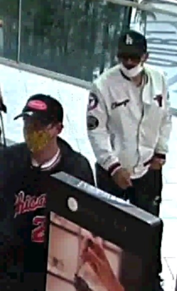 Two men in a mall. One man wearing a dark LA Dodgers baseball hat, white medical mask, white bomber jacket with a Chicago Bulls crest and black pants. The second male wearing a dark baseball hat with a red logo, a grey jacket and a dark shirt with the word Chicago and number 23 on the front of it.
