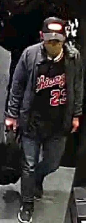 A man walking toward the camera, wearing a dark baseball hat with a red logo (the logo looks white as the light shines on it), a grey jacket and a dark shirt with the word Chicago and number 23 on the front of it, carrying a black back pack in his right hand.