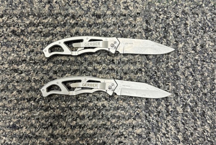 Two small silver folding knives with the word <q>Gerber</q> on the handle sit on a grey carpet