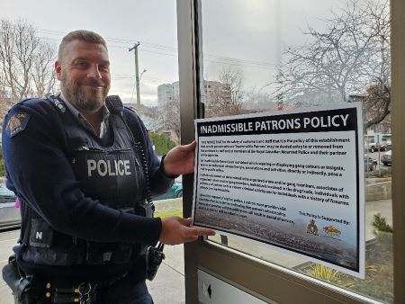 Un agent de police en uniforme tenant une affiche de la politique sur les clients admissibles devant la vitrine d’une entreprise.  